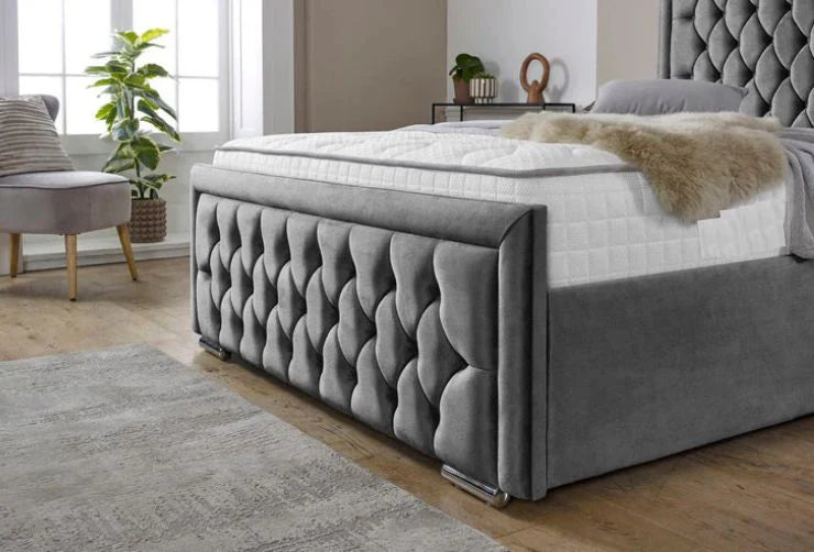 Gray tufted bed with white mattress in a room with a window and plants.