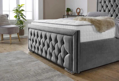Gray tufted bed with white mattress in a room with a window and plants.
